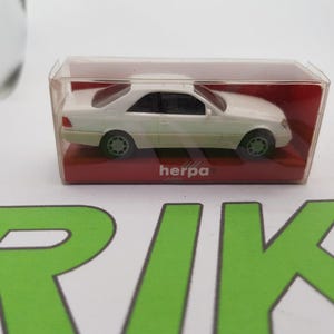 May include: White die-cast model car in a clear plastic display case with a red base. The car is a Mercedes-Benz and the case has the word "herpa" printed on it.