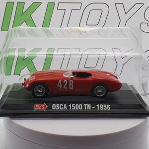 May include: A red 1:18 scale diecast model car of an OSCA 1500 TN from 1956. The car has a white number "428" on the side. The car is displayed in a clear plastic case on a black base with the words "OSCA 1500 TN - 1956" printed on it.