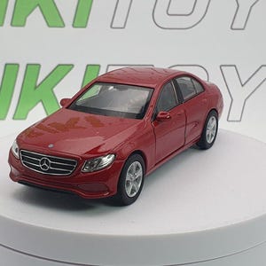 Puede incluir: Un coche modelo rojo, un sedán Mercedes-Benz, se exhibe sobre una plataforma blanca. El coche tiene ruedas plateadas y una parrilla delantera detallada. El fondo muestra la palabra "RIKI TOYS" en verde.