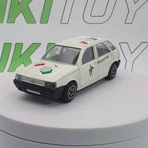 May include: A white die-cast model car with black wheels and trim. The car features decals with green, red, and white accents, and the text "Italia '90". The toy car is on a white surface.