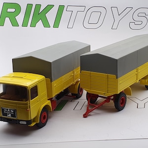 May include: A yellow toy truck with a gray canvas cover and a yellow trailer with a gray canvas cover. The truck has the text "MAN" and "WIESBADEN" on the side.