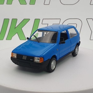 May include: A blue die-cast model car of a Fiat Uno. The miniature vehicle has a black front bumper, side mirrors, and window frames. The car is on a white surface with the word "RIKI TOYS" in green and white in the background.