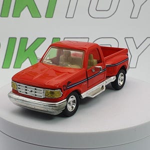 Può includere: Un camioncino giocattolo rosso con paraurti anteriore e pedane argentate. Il camion ha una striscia nera e blu lungo il lato e interni beige. Il giocattolo è su una superficie bianca.