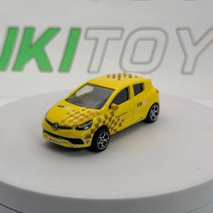 Può includere: Un'auto giocattolo Renault Clio gialla con ruote nere. L'auto presenta un motivo a scacchi sul davanti e la scritta "CLIO" sul lato. L'auto giocattolo è su una superficie bianca.