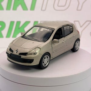 Può includere: Un'auto modello Renault Clio color beige. L'auto giocattolo ha finiture nere, ruote argentate e fari trasparenti. L'auto è esposta su una piattaforma rotante bianca. La parola "RIKI TOYS" è visibile sullo sfondo.
