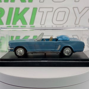 May include: A blue convertible toy car in a clear display case. The miniature car has silver wheels and a tan interior. The display case sits on a black base, and the background features the word "RIKI TOYS" in green and white.