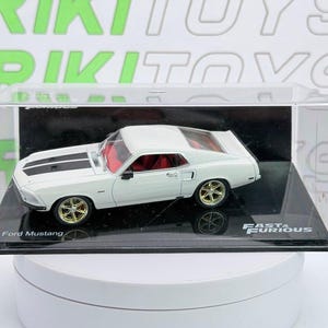 May include: A white Ford Mustang die-cast model car with black hood stripes, gold-colored wheels, and a red interior, enclosed in a clear plastic display case with a black base labeled Fast & Furious and Ford Mustang on the front panel.