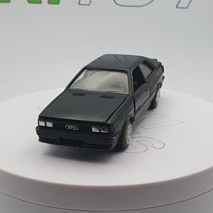 May include: A black toy car model of an Audi Quattro, a classic rally car. The car is in a slightly angled view, showing the front and side of the car. The car has black wheels and a black body.