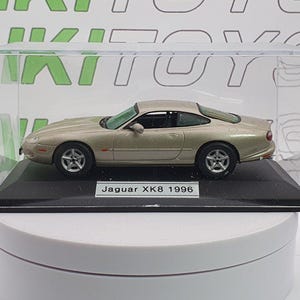 Può includere: Un'auto modello Jaguar XK8 1996 color oro, esposta in una teca di acrilico trasparente. Il modello è su una base nera con l'etichetta "Jaguar XK8 1996". L'auto ha ruote dettagliate e un design elegante.