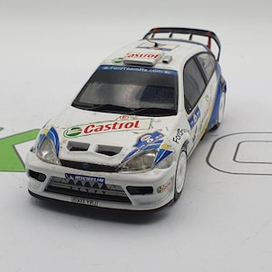 Könnte beinhalten: Weißes Diecast-Modell eines Ford Focus WRC Rallye-Autos mit Castrol- und FordTeamRS.com-Branding. Das Auto hat einen blauen und gelben Rennstreifen und einen schwarzen Michelin-Reifenaufkleber. Das Auto hat ein Nummernschild mit der Aufschrift "E003 YRJ".