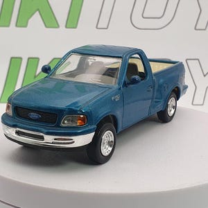 Può includere: Un giocattolo di camioncino Ford F-150 color teal. Il modello ha un paraurti anteriore argentato, pneumatici neri e interni dettagliati. Il logo Ford è visibile sulla parte anteriore. Il giocattolo è su una superficie bianca.