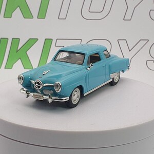 May include: A light blue diecast model of a vintage Studebaker Champion coupe. The miniature car features chrome accents, white-walled tyres, and a detailed interior. The model is displayed on a white rotating platform.