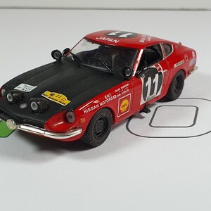 Può includere: Modello di auto da rally Nissan 240Z rosso e nero in scala 1:18 con il numero 11 sul lato. L'auto ha un cofano nero e un adesivo bianco "Nissan Motors" sul lato.