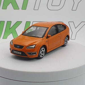 Peut inclure: Modèle réduit orange d'une Ford Focus ST. La voiture miniature a des roues noires avec des jantes argentées, une calandre noire et le texte "Focus ST" à l'avant. La voiture est sur une surface blanche avec un fond vert et blanc.