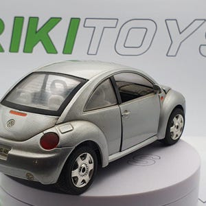 Peut inclure: Petite voiture argentée avec des pneus noirs et des feux arrière rouges. La voiture a une forme arrondie et un logo Volkswagen à l'arrière. Le mot "RIKI TOYS" est visible en vert en arrière-plan.