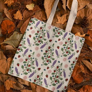May include: A cream-colored tote bag with a repeating pattern of strawberries, blueberries, lavender, and pink flowers. The bag has off-white handles and is set against a backdrop of autumn leaves.