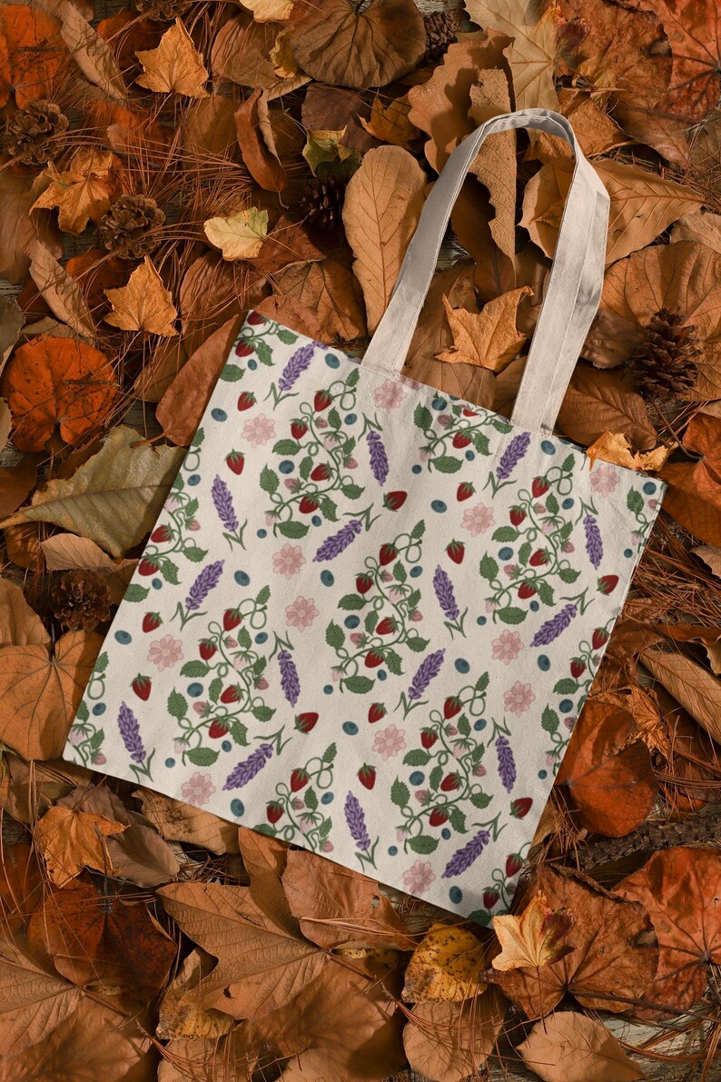 May include: A cream-colored tote bag with a repeating pattern of strawberries, blueberries, lavender, and pink flowers. The bag has off-white handles and is set against a backdrop of autumn leaves.