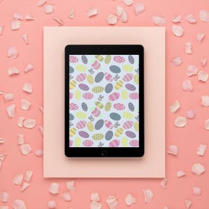 May include: A black tablet shows a colourful Easter egg pattern on a light blue background. The tablet is on a light pink rectangle, surrounded by a pink background and scattered flower petals. The egg design includes various colours and patterns.