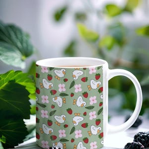 Puede incluir: Taza de cer&aacute;mica blanca con un patr&oacute;n repetido de patos de dibujos animados, fresas y flores rosas sobre un fondo verde claro. La taza tiene un asa curva y est&aacute; llena de un l&iacute;quido marr&oacute;n claro. Un producto de cocina y comedor.