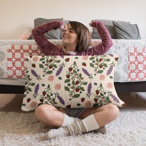 May include: Rectangular pillow with a cream background featuring a repeating pattern of strawberries, blueberries, lavender, and pink flowers. The pillow is held by a person sitting on a white rug.