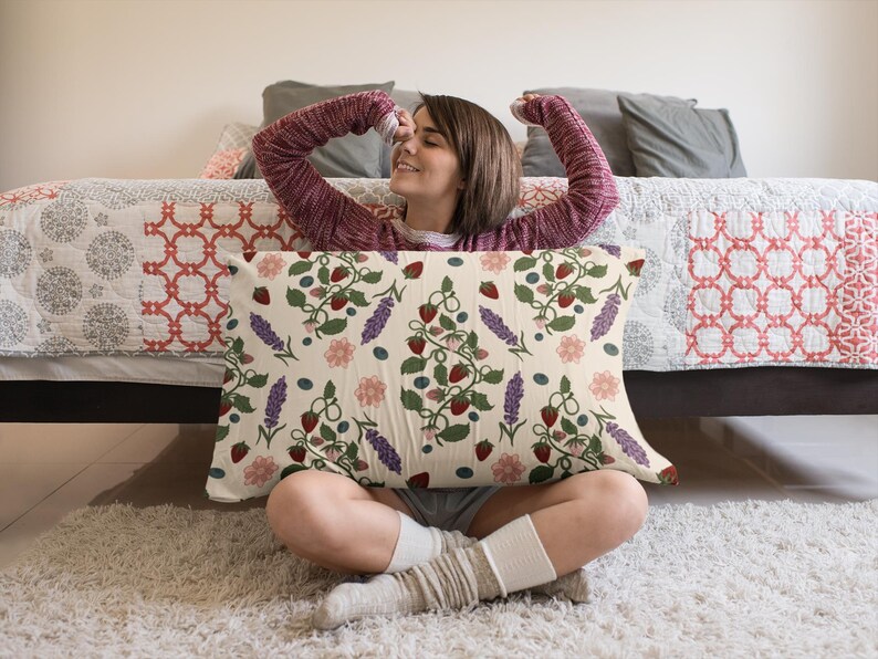 May include: Rectangular pillow with a cream background featuring a repeating pattern of strawberries, blueberries, lavender, and pink flowers. The pillow is held by a person sitting on a white rug.
