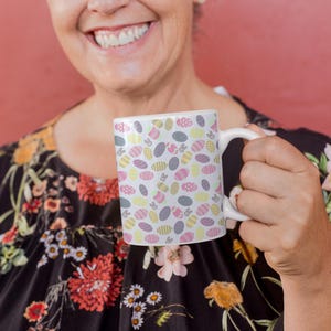 May include: White ceramic mug featuring a repeating pattern of Easter eggs in pastel shades. The eggs are decorated with various designs and colours, including pink, yellow, and grey. The mug has a white handle and is held by a person.