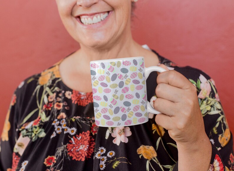 May include: White ceramic mug featuring a repeating pattern of Easter eggs in pastel shades. The eggs are decorated with various designs and colours, including pink, yellow, and grey. The mug has a white handle and is held by a person.