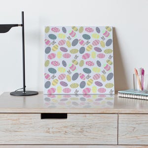 May include: A canvas print with a repeating pattern of Easter eggs and bunny silhouettes in pastel shades. It sits on a light wood dresser, alongside a black desk lamp, a black mug, and a glass of pens. The dresser is approximately 100 cm wide.