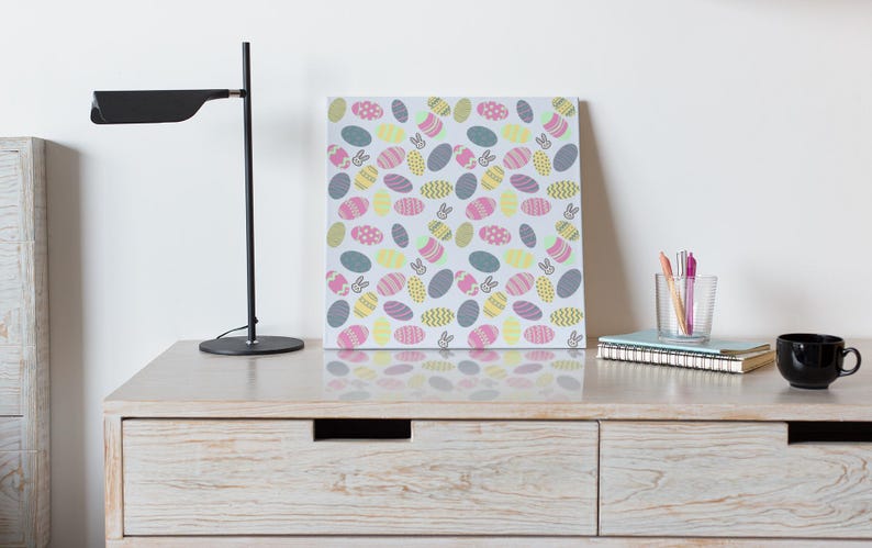 May include: A canvas print with a repeating pattern of Easter eggs and bunny silhouettes in pastel shades. It sits on a light wood dresser, alongside a black desk lamp, a black mug, and a glass of pens. The dresser is approximately 100 cm wide.