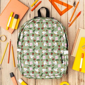 Puede incluir: Una mochila verde con un estampado de patos blancos, flores rosas y fresas rojas. Alrededor de la mochila hay &uacute;tiles escolares, como l&aacute;pices, ceras y un transportador, todo sobre una superficie de madera.