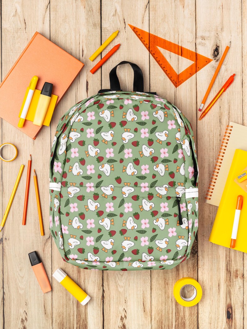 Puede incluir: Una mochila verde con un estampado de patos blancos, flores rosas y fresas rojas. Alrededor de la mochila hay &uacute;tiles escolares, como l&aacute;pices, ceras y un transportador, todo sobre una superficie de madera.