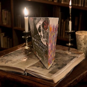 May include: A book titled "Haunting Adeline" with a skull on the cover. The book is standing upright on a wooden table with an open book in front of it. Two lit candles are on either side of the book.