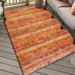 Könnte beinhalten: Ein Outdoor-Teppich in warmen Farben wie Orange, Rosa und Braun. Das Design zeigt ein Streifenmuster mit geometrischen Akzenten. Er liegt auf einer Holzterrasse, neben einem Korbmöbel und einer Topfpflanze.