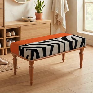 May include: A rectangular bench featuring a cushioned top with a bold black and white zebra stripe pattern on one side and a solid burnt orange section on the other. The bench is supported by four turned wooden legs with a light brown finish.