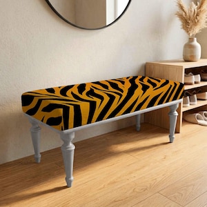 May include: A rectangular bench featuring a bold black and orange tiger stripe pattern on the upholstered seat. The bench is supported by four light gray, turned wooden legs that provide a stable base for the cushioned seating surface.