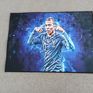 May include: A blue and black doormat with a photo of a soccer player in a blue jersey with the number 0 on the front. The player is making a gesture with his hands, as if he is blocking out noise.