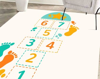 Puede incluir: Una alfombra de juego blanca con un diseño de rayuela con números naranja y azul verdoso del 1 al 7, una estrella y huellas.