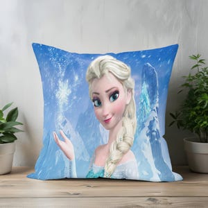 Op de afbeelding: Vierkant kussen met een cartoonafbeelding van Elsa uit Frozen. Het kussen heeft een blauwe achtergrond met een besneeuwde berglandschap. Elsa wordt afgebeeld met blond haar en een blauwe jurk.
