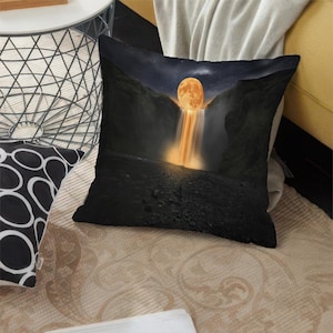 Pode incluir: Almofada quadrada decorativa com uma paisagem surrealista. O design retrata uma lua cheia laranja caindo em cascata de uma cachoeira entre penhascos escuros sob um céu estrelado. A almofada é provavelmente feita de tecido macio.