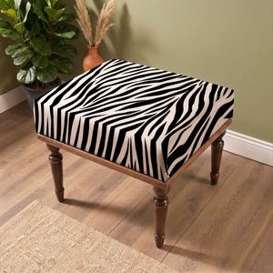 May include: A square ottoman featuring a bold black and white zebra stripe pattern on the upholstered top. The piece is supported by four dark brown wooden legs with a turned, tapered design that provides a stable base for the cushioned seat.