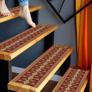 Puede incluir: Peldaños de escalera con un diseño estampado en tonos rojos, beige y dorado. Las huellas son rectangulares y se adaptan a escaleras de madera. El diseño presenta un motivo repetitivo similar al paisley. Las escaleras son de madera clara.