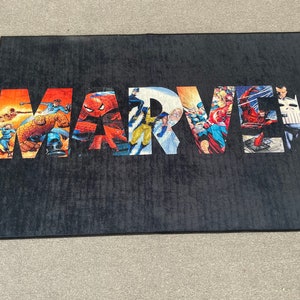 May include: Black rectangular rug featuring the word "MARVEL" in large, colorful letters. Each letter is filled with comic book illustrations of various Marvel characters, including Spider-Man and Captain America.