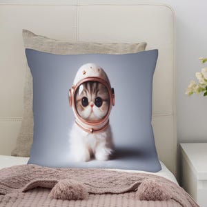 May include: A square decorative pillow featuring a cute kitten wearing a white and rose gold astronaut helmet. The kitten has large, expressive eyes and fluffy white fur. The pillow is on a bed with a gray headboard and a pink blanket.