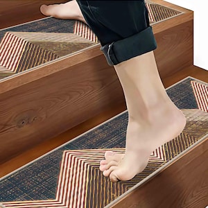 May include: Rectangular stair tread mat featuring a dark charcoal background with a geometric chevron pattern in white, tan, and gold lines. The text "STRONG GRIP, NON-SLIP" is printed at the top. The mat is installed on a wooden staircase.