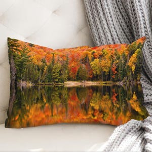 May include: Rectangular decorative pillow featuring a scenic autumn landscape. The image shows a vibrant forest with trees in shades of red, orange, and yellow reflected in a calm lake. The pillow is placed on a white surface with a gray knitted blanket.
