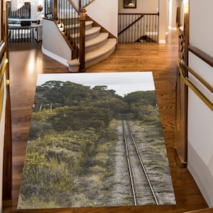 May include: A rectangular area rug featuring a scenic view of a railway track running through a lush, green landscape. The rug's design includes a detailed depiction of the tracks, vegetation, and a cloudy sky, creating a naturalistic scene.