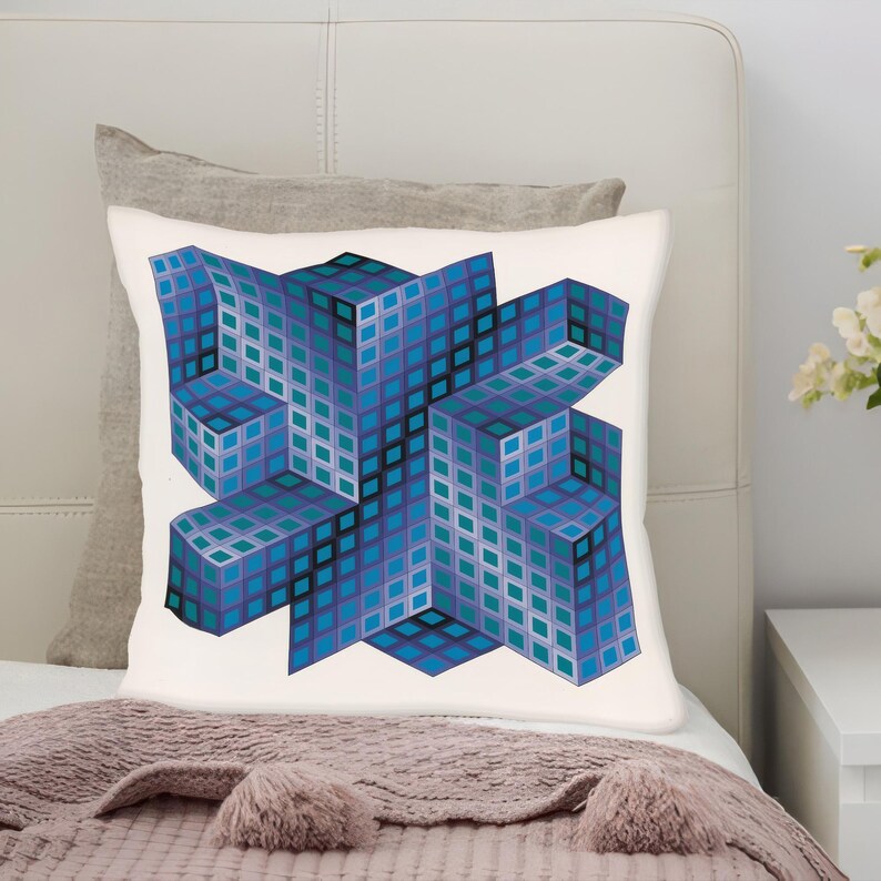 Op de afbeelding: Een vierkant wit kussen met een geometrisch ontwerp van blauwe, turquoise en paarse kubussen. De kubussen zijn in een driedimensionaal patroon gerangschikt, waardoor een optische illusie ontstaat. Het kussen ligt op een bed met een grijs kussen en een roze deken.