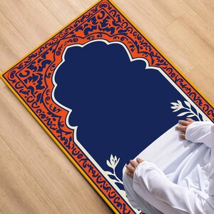 Peut inclure: Un tapis de prière avec un centre bleu marine et une bordure décorative orange et blanche. Le tapis présente un motif en arche traditionnel et est posé sur une surface en bois.
