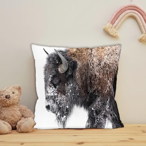 May include: Square throw pillow featuring a detailed image of a bison covered in snow. The bison is mostly black and brown with curved horns. The pillow is on a wooden surface with a teddy bear and a rainbow wall hanging in the background.