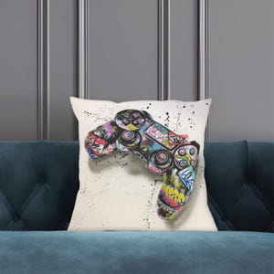 Op de afbeelding: Vierkantig kussen met een witte achtergrond, met een graffiti-achtige afbeelding van een gamecontroller. Het controllerontwerp is veelkleurig met zwarte verfspatten. Het kussen ligt op een blauwe bank.